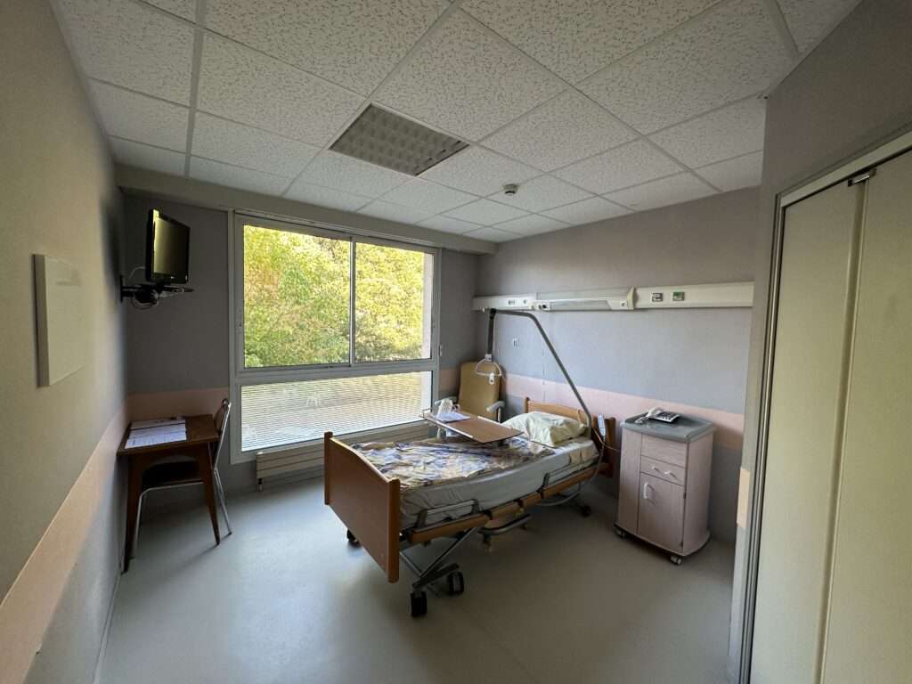 Chambre de l'hôpital, lit médicalisé avec une table de chevet sur le côté ainsi qu'une télévision accrochée au mur face au lit. Il y a une grande fenêtre donnant sur la cour.